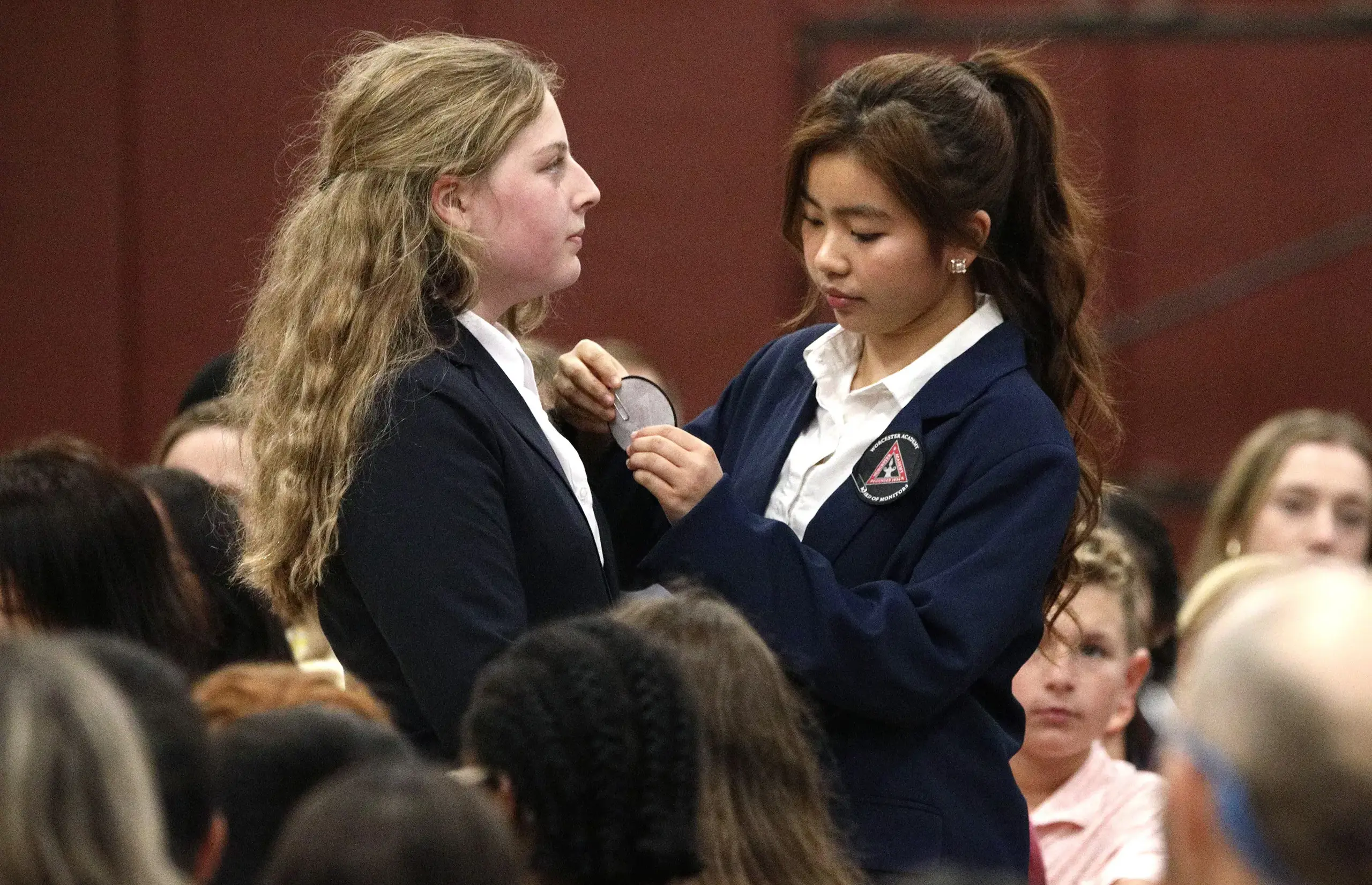 Two girls in school uniforms standing in a crowd. One is pinning an award onto the other's jacket.